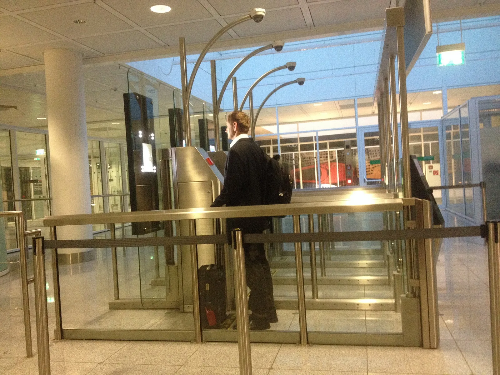 A traveller with a backpack and a wheeled case passing through an automated border gate, seen from behind