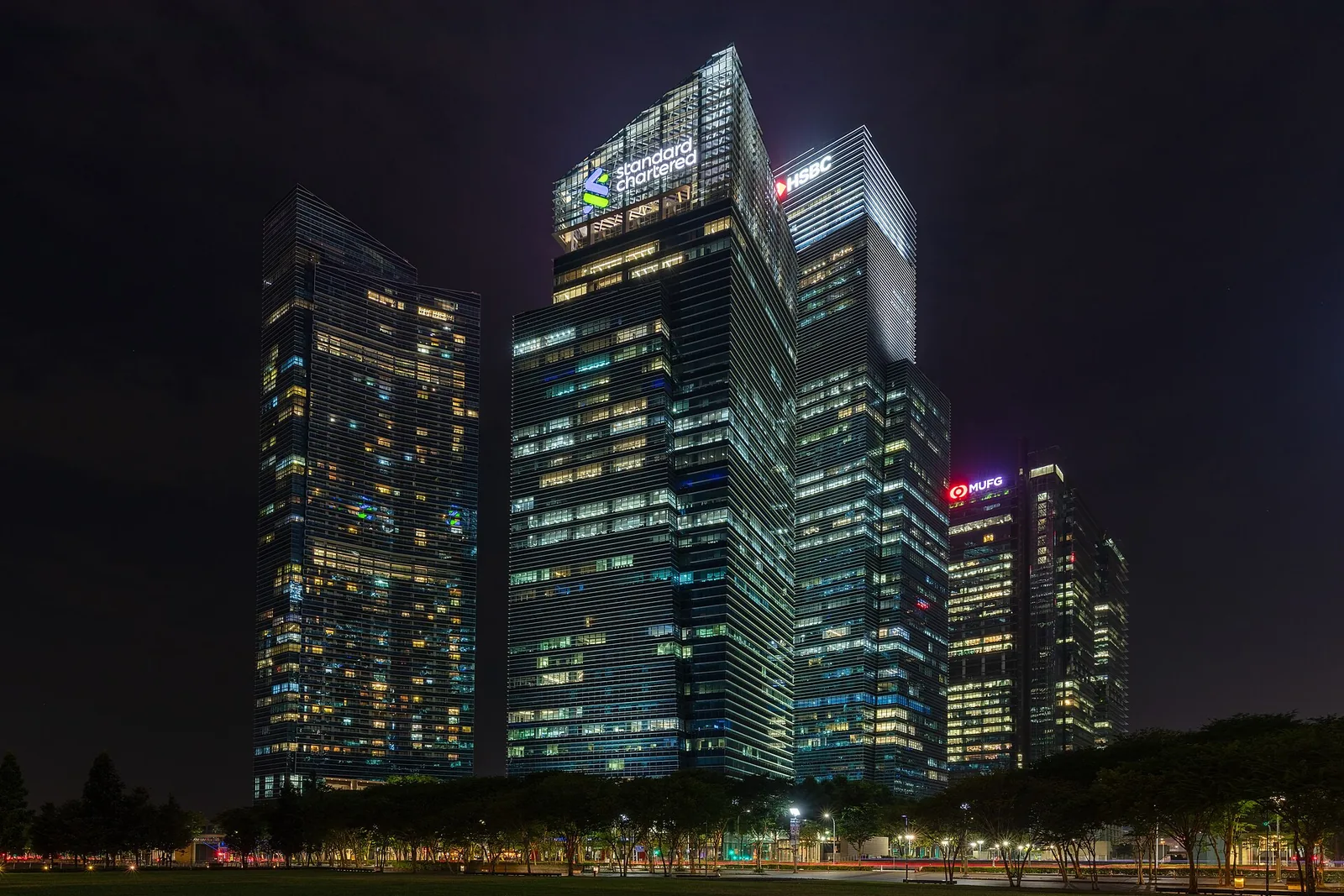 Glass office towers in Singapore's Marina Bay financial district lit up at night, with trees and a lawn in the foreground