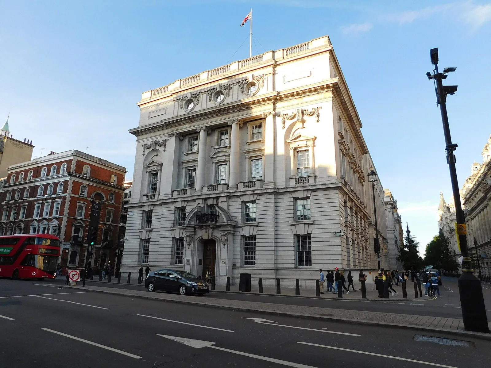 A stone government building on a corner of Whitehall in London, a red double-decker bus passing on the road outside