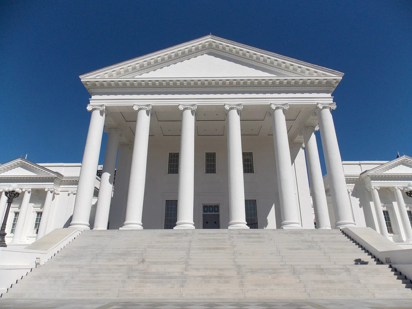 The white temple-front portico of the Virginia State Capitol in Richmond — Ionic columns beneath a pediment, broad steps in front, flanking wings to either side under a clear sky